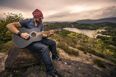 Mann mit Mütze spielt akustische Gitarre auf einem Felsen über einer Seenlandschaft