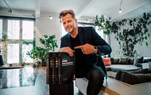 Alex Fischer zeigt in einer Loft-Wohnung auf eine Box mit seinen Kursunterlagen
