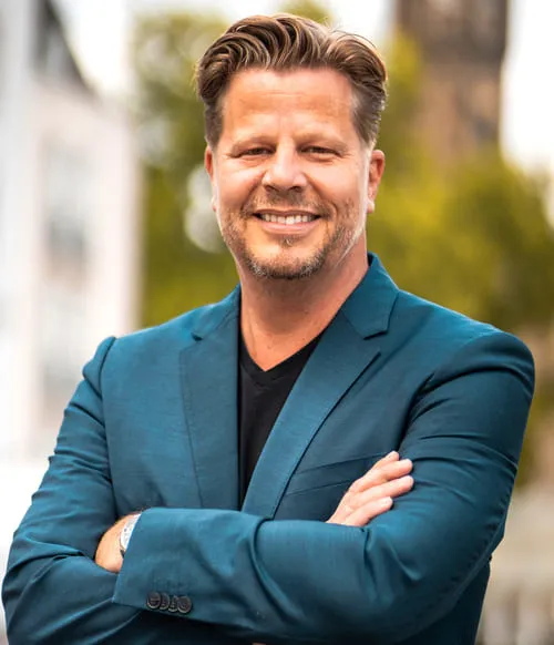 Lächelnder Alex Fischer im grünen Sakko mit verschränkten Armen vor unscharfem Stadthintergrund