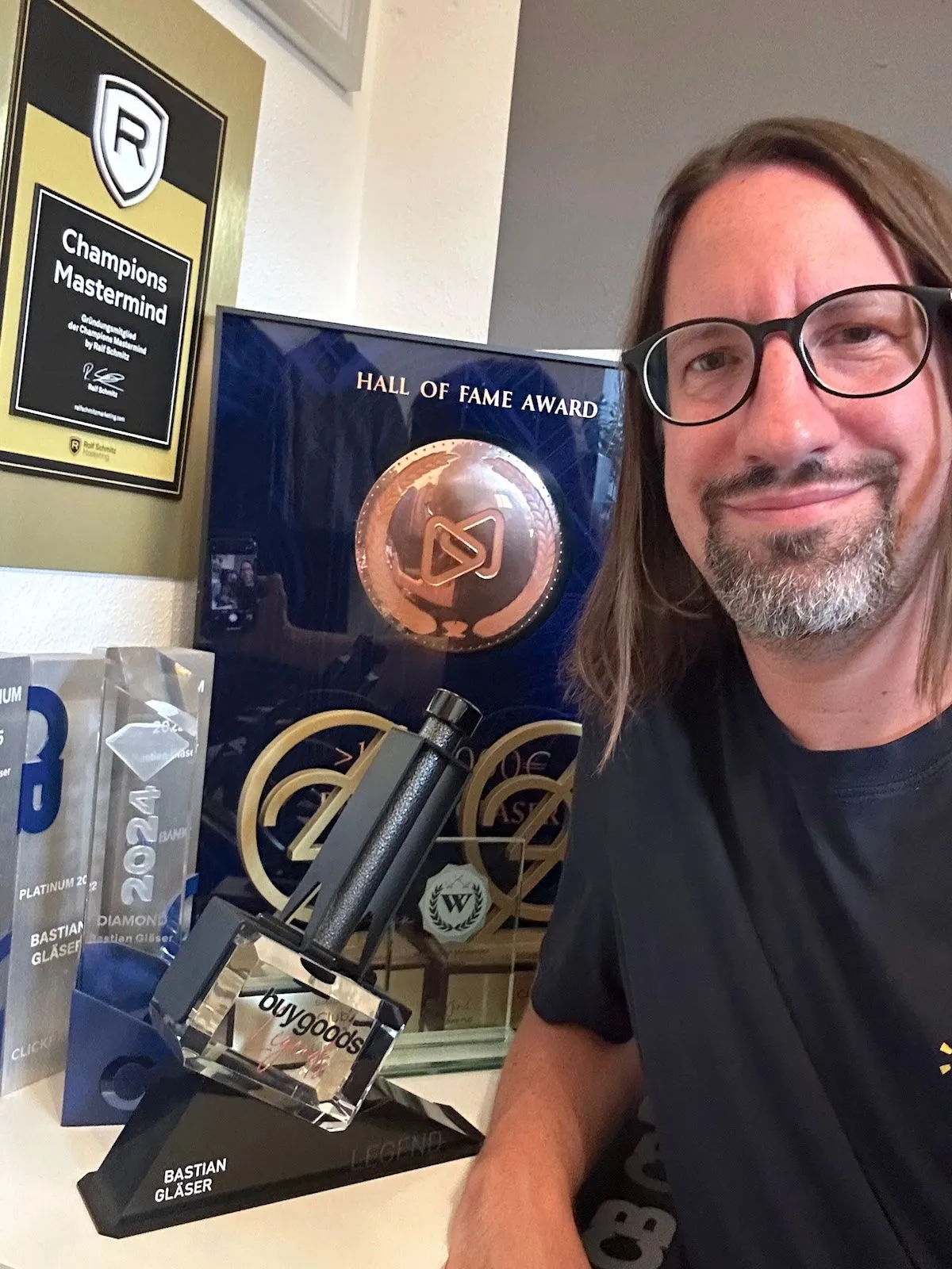 Bastian Gläser mit Brille und langen Haaren lächelt in die Kamera, im Hintergrund Auszeichnungen im Regal