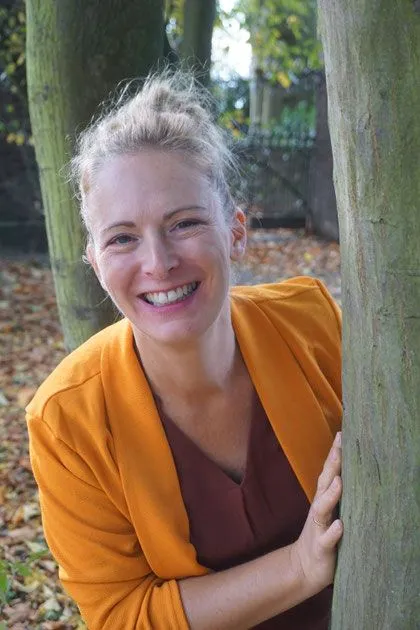 Porträt von Carina Mathes im Herbst, lächelnd an einen Baumstamm gelehnt