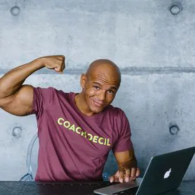 Coach Cecil im Coach-Cecil-Shirt spannt den Bizeps an und sitzt vor einem Laptop