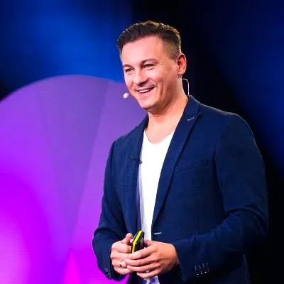 Dawid Przybylski im dunkelblauen Sakko lacht mit Headset auf einer Bühne in violettem Licht
