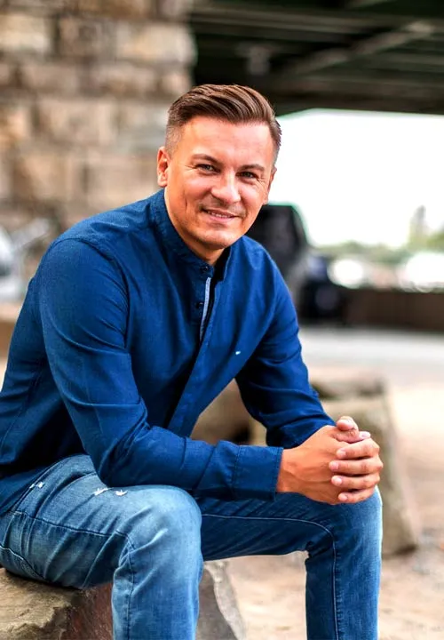 Dawid Przybylski im blauen Pullover sitzt lächelnd auf einer Steinmauer