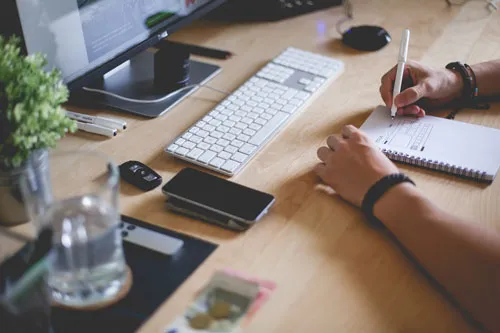 Schreibtisch mit Monitor, Tastatur, Smartphone und einer Person, die sich Notizen macht – Symbolbild zum Ecom Launch System