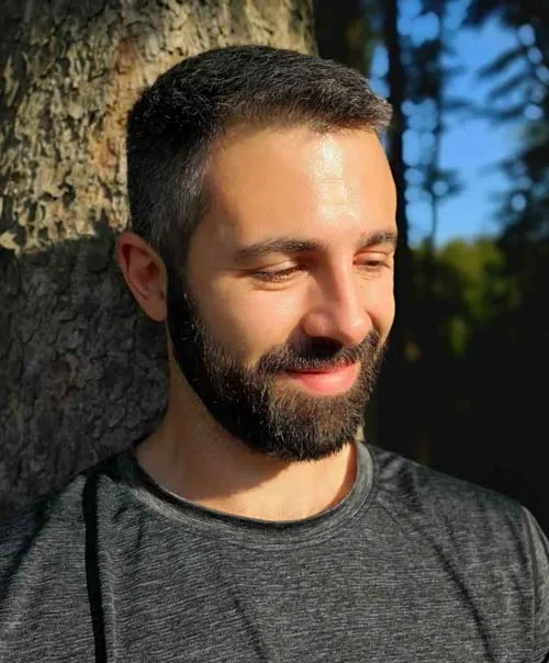 Eric Hüther mit Bart im grauen T-Shirt vor einem Baum in der Sonne