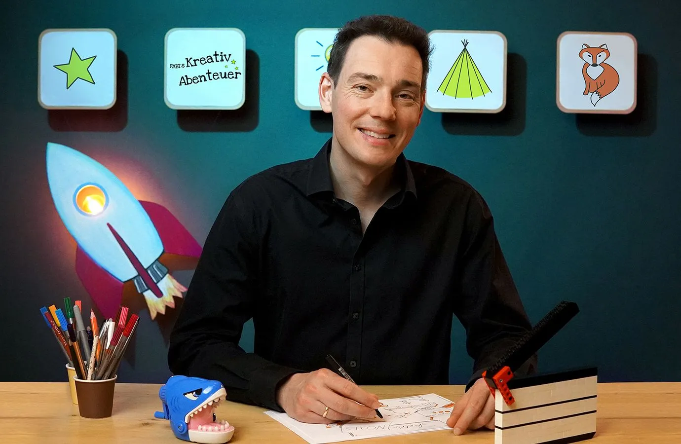 Fabian Kreis sitzt lächelnd an einem Zeichentisch mit Stiften, hinter ihm eine Wand mit Rakete und Bildkacheln