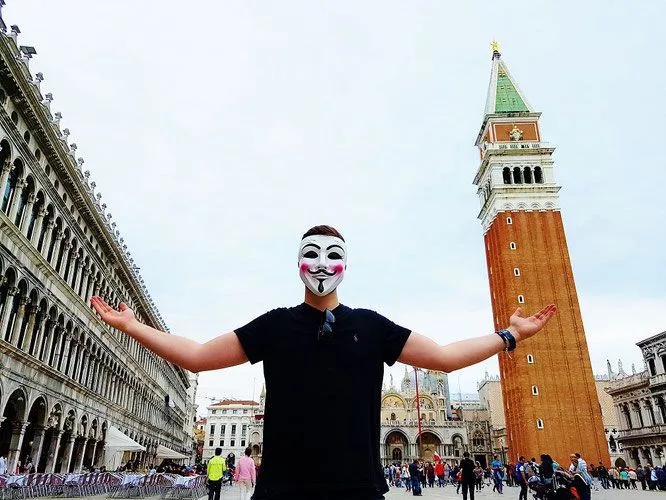 Flo Pharell mit weißer Maske und ausgebreiteten Armen vor dem Campanile auf dem Markusplatz in Venedig