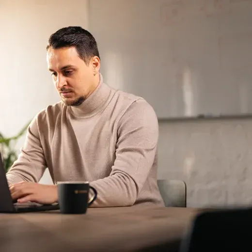 Florian Schäfer im hellen Rollkragenpullover am Laptop mit einer Kaffeetasse