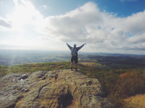 Wanderer steht mit ausgebreiteten Armen auf einem Felsen über einer weiten Landschaft – Inspire your Life