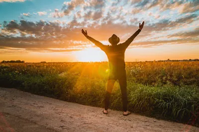 Mann mit Hut breitet vor einem Sonnenuntergang über einem Feld die Arme aus – Symbolbild für Erfahrungen mit dem Lebensstarter