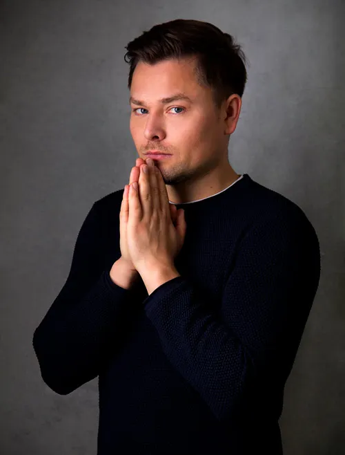 Maxim Mankevich im schwarzen Pullover mit aneinandergelegten Händen vor grauem Hintergrund