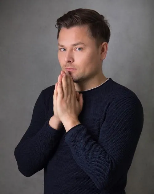 Maxim Mankevich im dunklen Pullover mit gefalteten Händen vor grauem Hintergrund