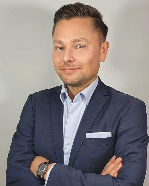 Maxim Mankevich im dunkelblauen Anzug mit verschränkten Armen vor grauem Hintergrund