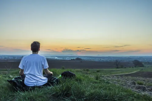 Mann meditiert auf einer Wiese mit Blick auf den Sonnenuntergang – Symbolbild für Erfahrungen mit der Meditation Mastery