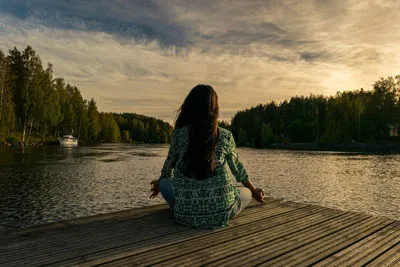 Frau meditiert im Schneidersitz auf einem Steg am See bei Sonnenuntergang – Symbolbild zur Meditation Mastery