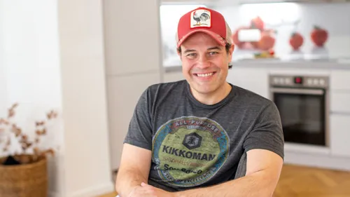 Mike Hager mit rotem Cap und Kikkoman-T-Shirt sitzt lächelnd in einer Küche