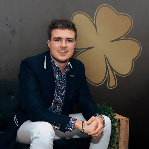 Nick Geringer sitzt im dunklen Sakko vor einer Wand mit goldenem Kleeblatt