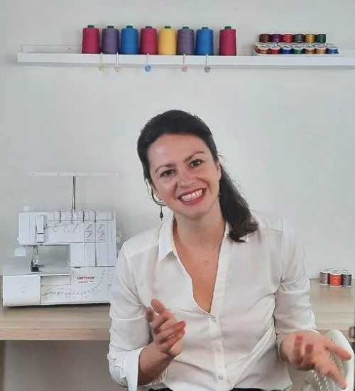 Nicole Langer lächelt vor ihrer Overlock-Nähmaschine und einem Regal mit bunten Garnrollen