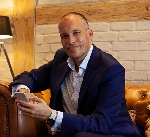 Prof. Dr. Oliver Pott lächelt im Anzug auf einem braunen Ledersofa mit Smartphone in der Hand
