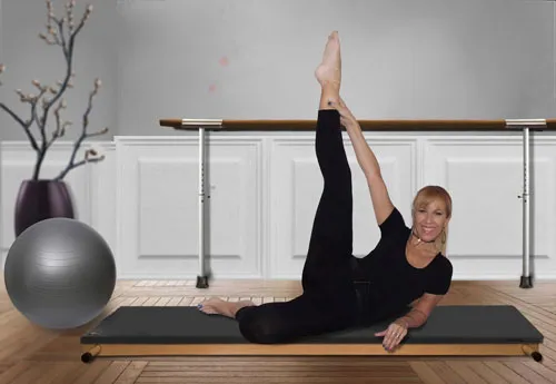 Pilates-Trainerin macht eine seitliche Beinhebe-Übung auf einer Matte vor einer Ballettstange