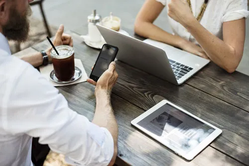 Zwei Personen mit Laptop, Smartphone und Tablet an einem Holztisch im Café