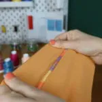 Hände halten ein orangefarbenes, mit der Overlock genähtes Stoffstück mit bunter Ziernaht