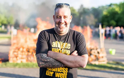 Ralf Schmitz mit verschränkten Armen im Affiliate-Marketing-T-Shirt vor einem Lagerfeuer