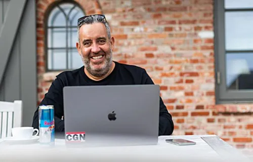 Ralf Schmitz mit Sonnenbrille im Haar sitzt lächelnd am Laptop vor einer Backsteinwand