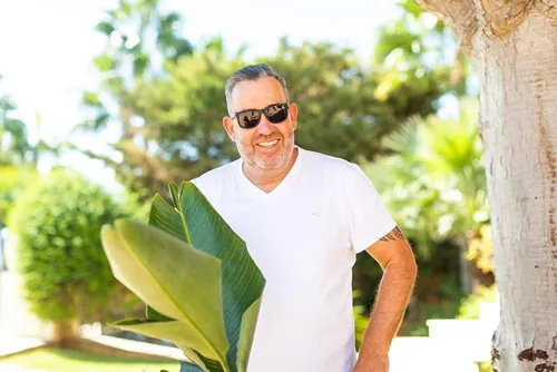 Ralf Schmitz im weißen T-Shirt mit Sonnenbrille lehnt zwischen Palmen und grünen Blättern