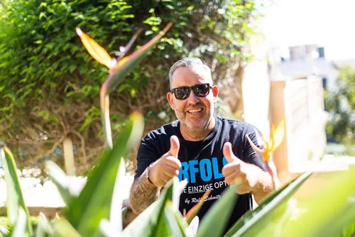 Ralf Schmitz im schwarzen T-Shirt zeigt zwischen Paradiesvogelblumen beide Daumen nach oben
