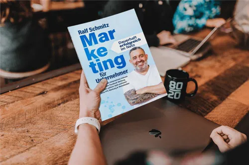 Hand hält das Buch Marketing für Unternehmer von Ralf Schmitz über einem Holztisch mit Laptop und Tasse
