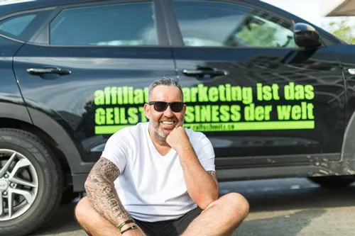 Ralf Schmitz sitzt vor einem Auto mit der Aufschrift „Affiliate-Marketing ist das geilste Business der Welt“
