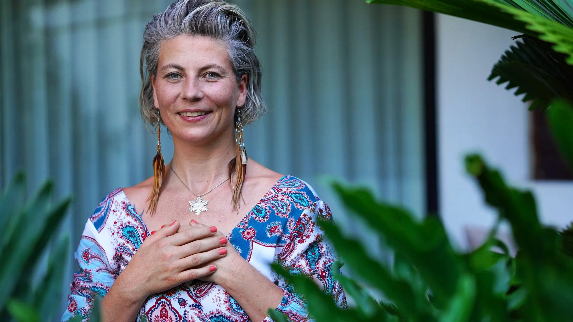 Sandra Hirsch mit grauen, zurückgesteckten Haaren, Federohrringen und Schneeflockenkette, die Hände auf der Brust, vor grünen Pflanzen
