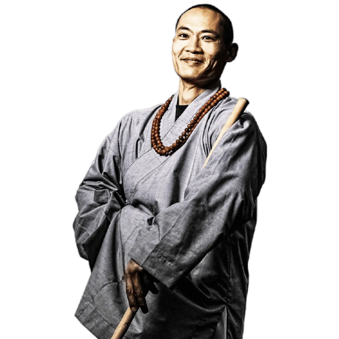 Shi Heng Yi in grauer Robe mit Gebetskette und einem Holzstab, lächelnd