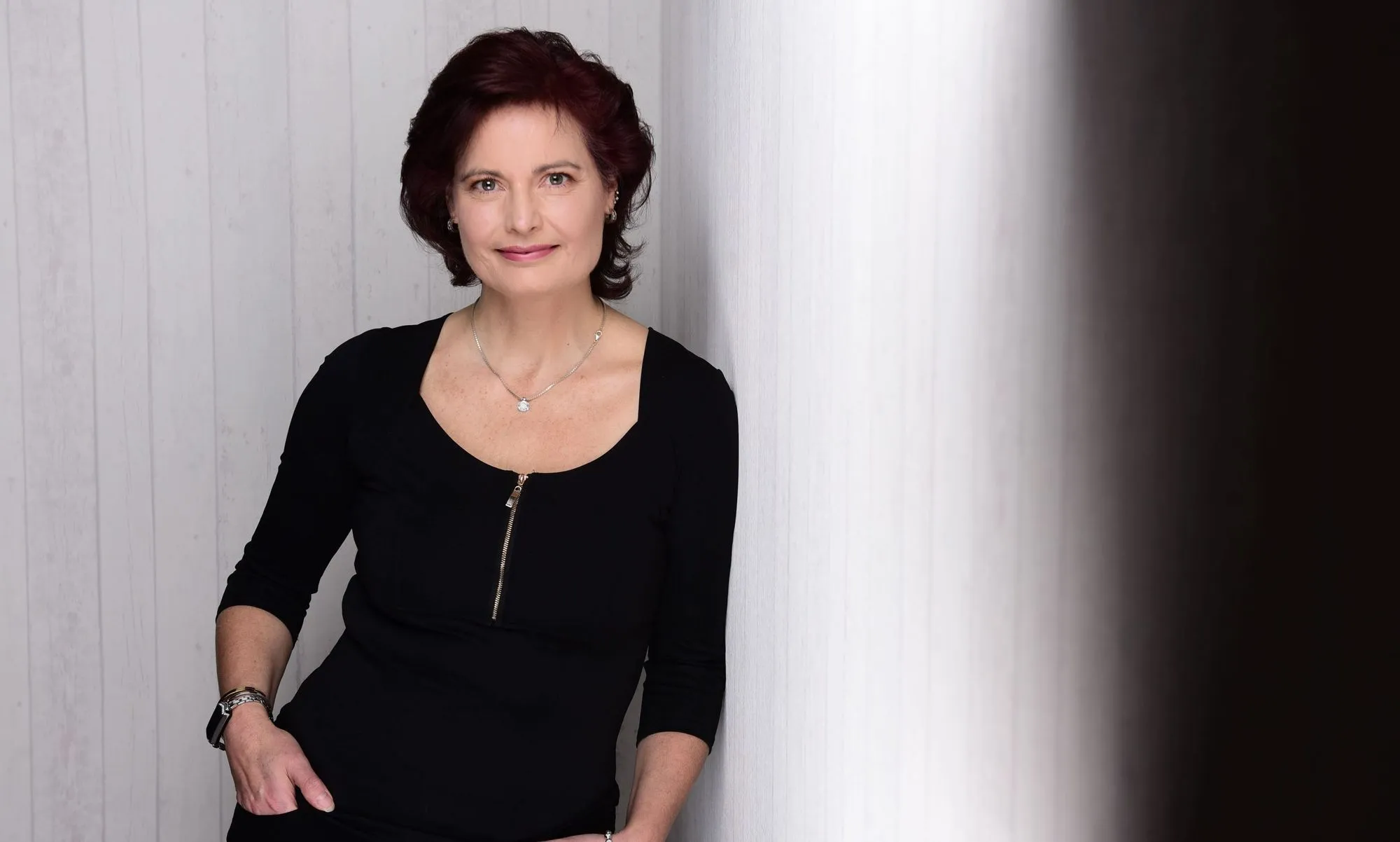 Dr. Susanna Wallis lehnt in schwarzem Oberteil an einer weißen Holzwand