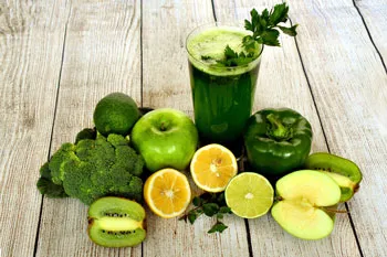 Grüner Smoothie im Glas umgeben von Äpfeln, Brokkoli, Kiwis, Zitrusfrüchten und Paprika